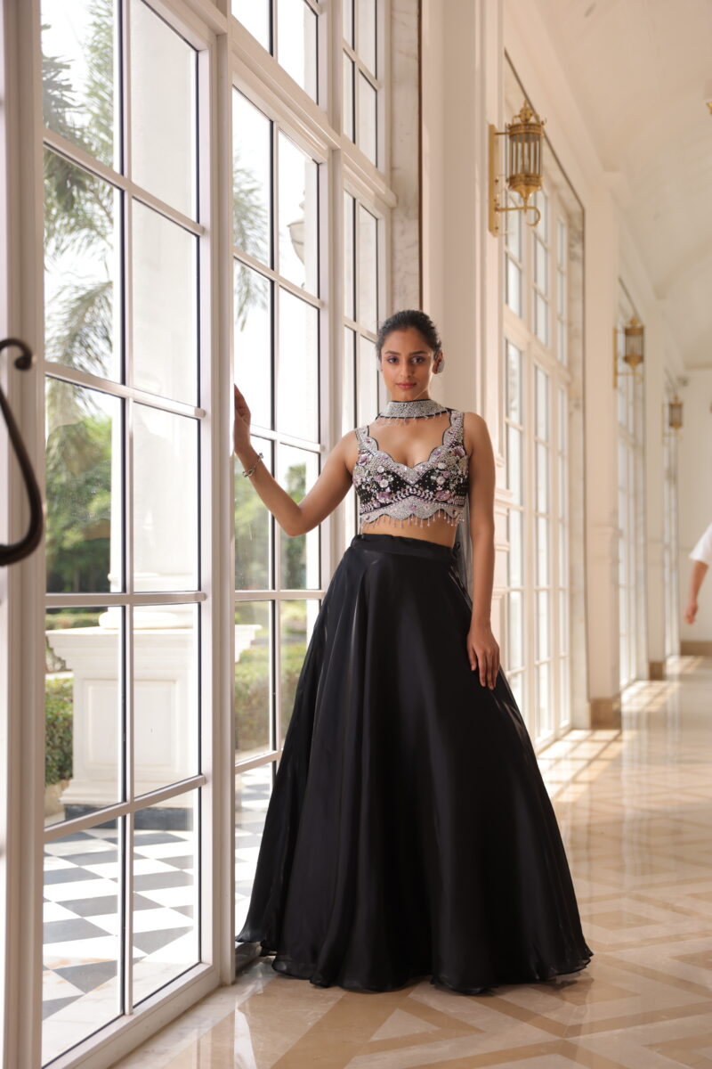 A model is dressed in a black stone studded embroidered lehenga, standing near a big door-sized window. - By Dishu Sarees (Lehenga to Buy Online) https://dishusarees.com/