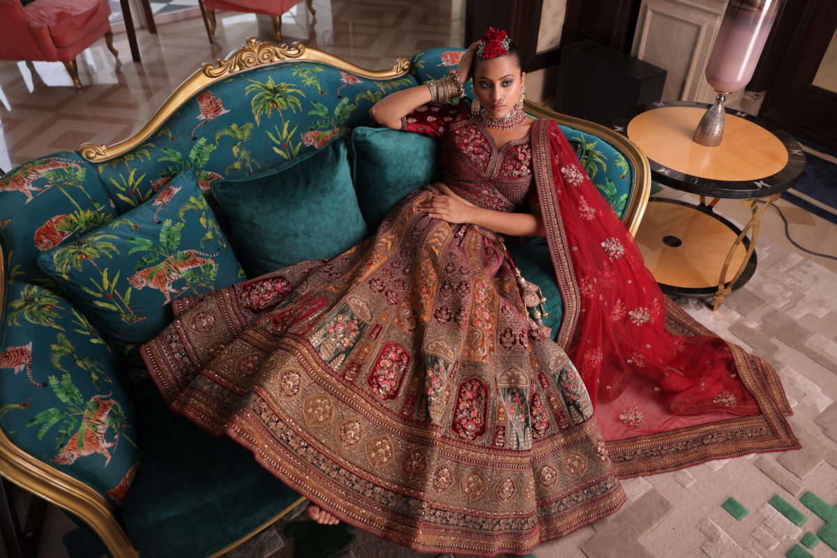 A model lying on a royal and rich fabric sofa dressed in bright red bridal lehenga. - By Dishu Sarees (Bridal Lehenga to Buy Online). https://dishusarees.com/