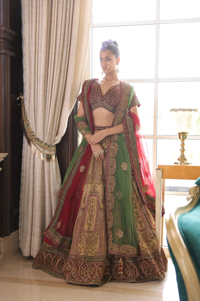 A model is standing near a door-sized window dressed in red-green bridal lehenga. By Dishu Sarees (Bridal lehenga to Buy Online).
https://dishusarees.com/