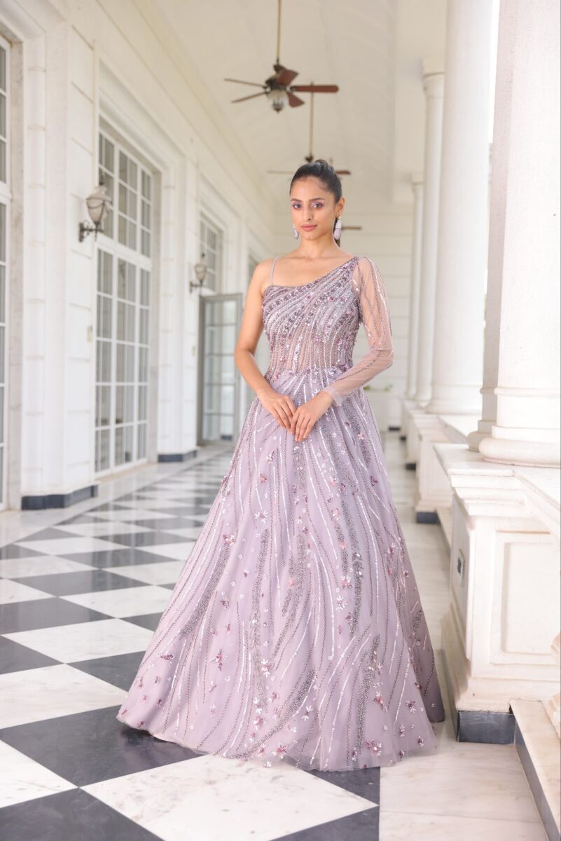 A model is wearing one shoulder lavender pink gown with shiny minimalist embroidery. She is standing in a white corridor. By Dishu Sarees (https://dishusarees.com/) Party Wear Gowns for Women.