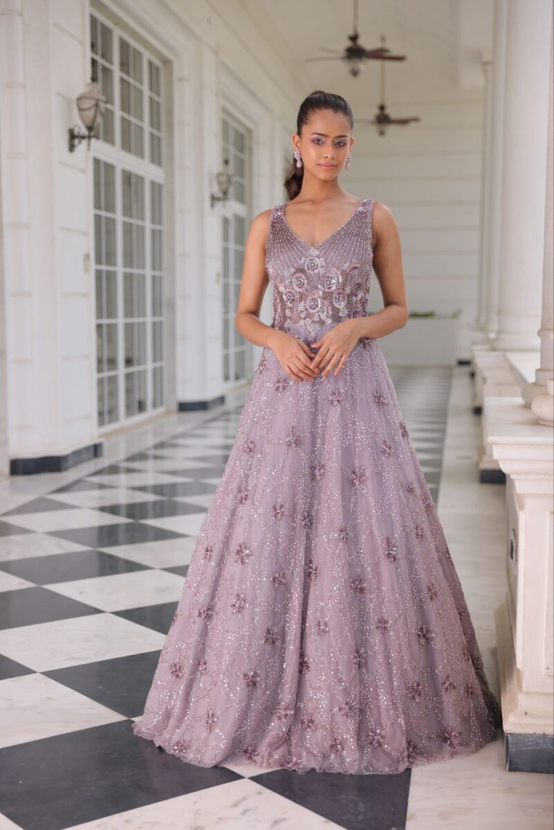 A model is wearing lavender pink floor length gown with shiny floral embroidery. She is standing in a white coloured corridor. - By Dishu Sarees (https://dishusarees.com/) Party Wear Gowns for Women.