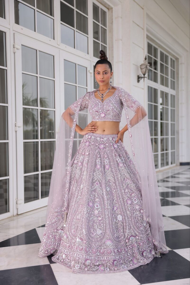 A model is wearing a lavender lehenga with detailed floral and embellished embroidery, and she is standing near a white door. - By Dishu Sarees (Designer Lehenga Buy Online) https://dishusarees.com/