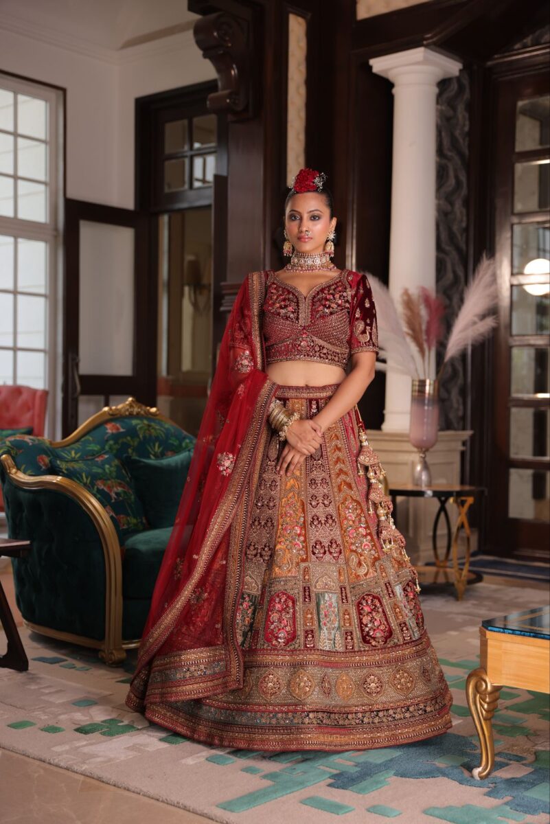 A model is wearing a red lehenga with dupatta that has beautiful Indian embroidery and zari work. She is standing in a vintagish living room.  - By Dishu Sarees (Designer Lehenga Buy Online) https://dishusarees.com/